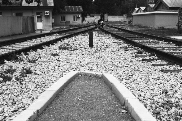 Lucky Lucky SHD 400 (FF)

English Alt Text:
A black-and-white photograph taken from a low angle, showing a railway junction where tracks split in two directions. The foreground features a gravel path bordered by concrete edges. White gravel surrounds the tracks. On both sides are small buildings, including houses and utility structures. In the distance, a person walks along the tracks, adding a human element to the otherwise industrial scene. The composition highlights perspective and contrast …