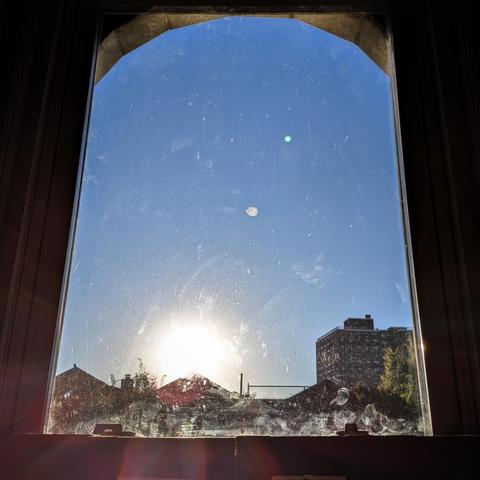 Looking through an arched window fifty-one minutes after sunrise the sun is just above the buildings in the blue sky, illuminating all of the dirt and scratches on the glass, and making a lens flare with multi-colored petals on the bottom left. Pointed roofs of Harlem brownstones are silhouetted across the street, and a taller apartment building can be seen in the distance.