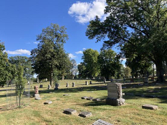 A serene cemetery scene featuring gravestones amidst green grass and trees, under a clear blue sky with scattered clouds.