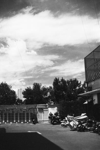 Lucky Lucky SHD 400 (FF)

English Alt Text:
A black-and-white image of an outdoor urban area. Several motorcycles and scooters are parked in the foreground, some covered with protective sheets. To the right is a building with a tiled roof. Trees and a tall building are visible in the background. The sky is cloudy, and faint vertical lines suggest film damage or scanning artifacts.

中文替代文字：
黑白照片描绘一个户外城市场景。前景停放着几辆摩托车和电动车，有些覆盖着防护布。右侧是一栋带瓦屋顶的建筑。背景有树木和一栋高楼。天空多云，画面上有些垂直线条，可能是胶片损伤或扫描痕迹。