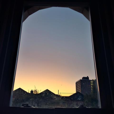 Looking through an arched window twenty-one minutes before sunrise the cloudless sky fades from light yellow at the horizon to dusty blue up above, with a band of pink in the lower sky. Pointed roofs of Harlem brownstones are silhouetted across the street, and a taller apartment building can be seen in the distance.