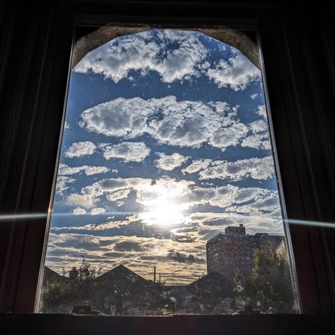 Looking through an arched window ninety minutes after sunrise the blue sky is filled with bands of complex, chunky, white clouds, with an orange tinge at the horizon. Pointed roofs of Harlem brownstones are silhouetted across the street, and a taller apartment building can be seen in the distance. The window is grimy on the bottom.