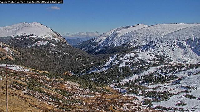 Scenic view from the official web cam at the Alpine Visitor Center in Rocky Mountain National Park.
Looking east from the visitor center on Trail Ridge Road, down into the valley below. A light covering of snow on the mountains across the valley, bright in full sunlight, a “bluebird sky” across the top. Between the two mountains can be seen cumulus clouds out over the flat steppe to the east. October 2025.