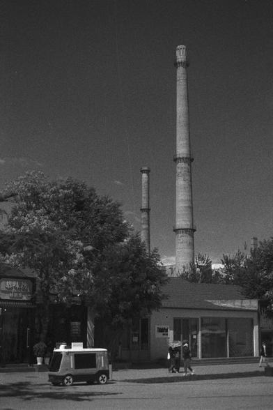 Ilford HP5 Plus 400 (FF)

English Alt Text:
A vintage-style black-and-white photo showing a city street with two tall industrial chimneys in the background, suggesting a factory or power plant. In the foreground, a small building with signage is partially hidden by trees. A small vehicle, possibly a delivery cart, is parked nearby. Two people walk along the street. The image contrasts urban life with industrial infrastructure, capturing a moment of quiet coexistence between human activity and t…