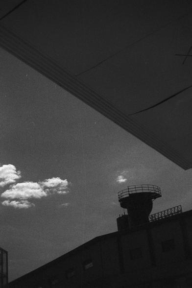 Ilford HP5 Plus 400 (FF)

English Alt Text:
A dramatic black-and-white photo taken from a low angle, showing a silhouetted building with a cylindrical tower on top, possibly a water tank or observation deck. The tower has a circular railing. Above, a large overhanging structure frames the top of the image, casting shadows. The sky is visible with a few clouds, contrasting with the dark foreground. The composition uses strong shadows and angles to create depth and mystery, evoking a sense of ind…