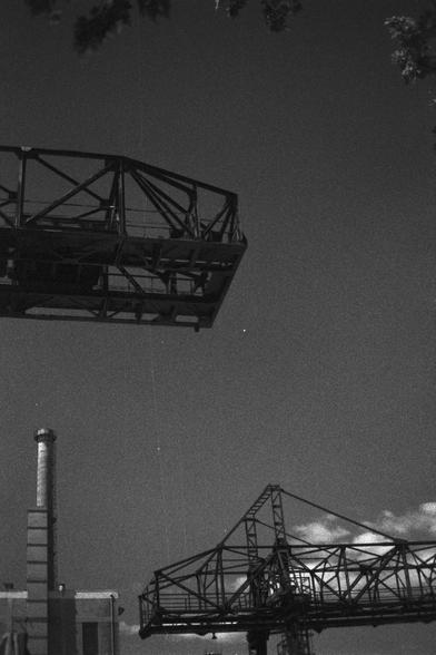 Ilford HP5 Plus 400 (FF)

English Alt Text:
A black-and-white photograph of an industrial landscape. Dominating the scene are two large metal structures resembling cranes or bridge segments, with intricate beams and frameworks. One structure is cropped in the upper left, while the other anchors the lower right. In the background stands a tall chimney attached to a factory-like building. The sky is mostly clear with a few scattered clouds. The image emphasizes geometric shapes and contrasts betw…