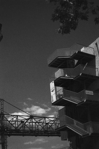 Ilford HP5 Plus 400 (FF)

English Alt Text:
A monochrome photo of a modern industrial building with angular balconies or staircases jutting outward. The structure features metal railings and a geometric design. A person is visible on one of the upper levels, and a sign is mounted on a balcony. In the background, a metal bridge or walkway connects parts of the building. A tree branch enters the frame from the upper right. The image captures a blend of human presence and architectural precision, …
