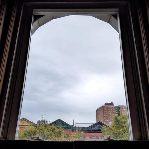 Looking through an arched window half an hour after sunrise the sky is full of blue-grey rain clouds. Pointed roofs of Harlem brownstones with red brickwork are across the street, and a taller apartment building can be seen in the distance. The green tops of two trees are on the bottom and right.