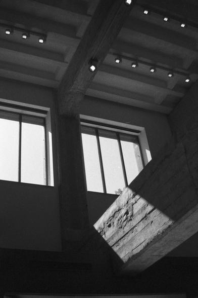 Ilford HP5 Plus 400 (FF)

English Alt Text:
A black-and-white photograph of a modern architectural interior. Tall vertical windows with metal frames allow sunlight to stream in, casting sharp shadows on rough concrete walls and beams. The space feels industrial and geometric, with strong lines and textures. Recessed ceiling lights are arranged in neat rows, adding to the clean, structured aesthetic. The interplay of light and shadow emphasizes the raw materials and creates a dramatic, contempla…