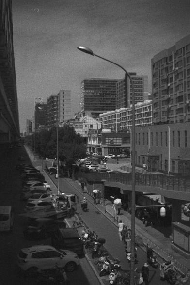 Ilford HP5 Plus 400 (FF)

English Alt Text:
A black-and-white photograph of a bustling city street. Mid-rise buildings with balconies line the right side, while an elevated structure casts shadows on the left. Cars, scooters, and pedestrians fill the scene, some carrying umbrellas. A tall streetlamp stands prominently in the foreground. The image captures the energy and density of urban life, with layered architecture and movement throughout.

中文替代文字：
这张黑白照片描绘了一个繁忙的城市街景。右侧是一排带阳台的中高层建筑，左侧则有一座高架结…