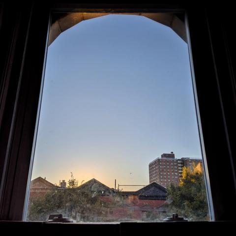 Looking through an arched window half an hour after sunrise the pale blue sky is cloudless, with a yellow glow at the horizon. Pointed roofs of Harlem brownstones with red brickwork are across the street, and a taller apartment building can be seen in the distance. The green tops of two trees are on the bottom and right.
