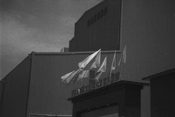 Ilford HP5 Plus 400 (FF)

English Alt Text:
A black-and-white image of a large industrial building with corrugated metal siding. Several flags mounted on poles flutter in the wind in front of the building. Chinese characters are visible on the upper facade and above the entrance, though partially obscured. The sky is clear, and the lighting suggests daytime. The image has a documentary feel, emphasizing the structure’s utilitarian design and the presence of national or institutional identity th…