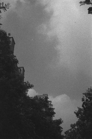 Ilford HP5 Plus 400 (FF)
English Alt Text:
A black-and-white photograph capturing a dramatic sky filled with textured clouds. On the left, partially hidden by dense tree foliage, are several multi-story buildings with rectangular windows—likely residential. Trees frame both sides of the image, creating a natural border. The sky dominates the scene with varying shades of gray, suggesting depth and movement. The grainy texture and lighting evoke a vintage, film-like aesthetic. The image feels int…