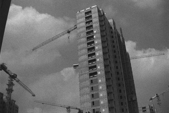 Ilford HP5 Plus 400 (FF)

English Alt Text:
A black-and-white photo of a high-rise building under construction. The structure is mostly complete, with its framework and exterior visible. Multiple tower cranes surround the building, indicating active construction. The sky is partly cloudy, and the image has a grainy texture that adds to its industrial and dramatic tone. The scene conveys the scale and complexity of urban development, with towering machinery and architectural ambition.

中文替代文字：
这…