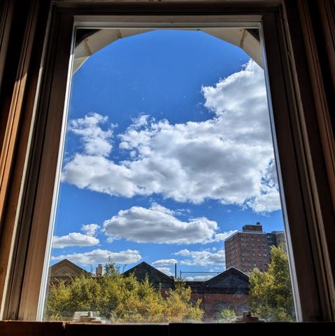 Looking through an arched window just before noon the bright blue sky is filled with large puffy  white clouds. Pointed roofs of Harlem brownstones are across the street, and a taller apartment building can be seen in the distance. The green tops of two trees are on the bottom and right.