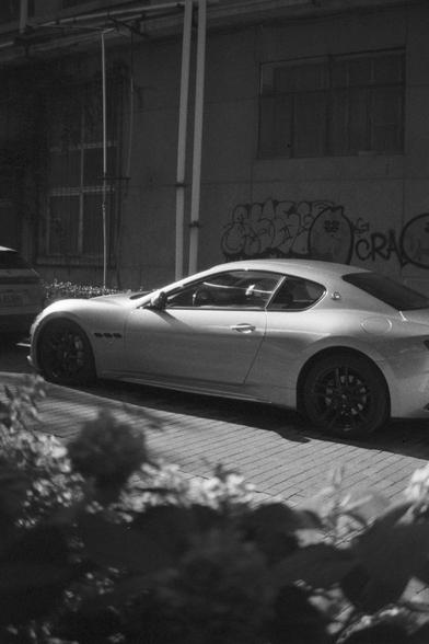 Ilford HP5 Plus 400 (FF)

English Alt Text:
A silver Maserati sports car is parked on a brick-paved street beside a wall covered in graffiti. The car has sleek curves, black alloy wheels, and a modern design. The photo is in black and white, giving it a timeless, cinematic feel. Blurred foliage in the foreground adds depth. Another vehicle is partially visible nearby. The contrast between the luxury car and gritty urban surroundings creates a visually compelling scene.

中文替代文字：
一辆银色玛莎拉蒂跑车停在砖铺街道…