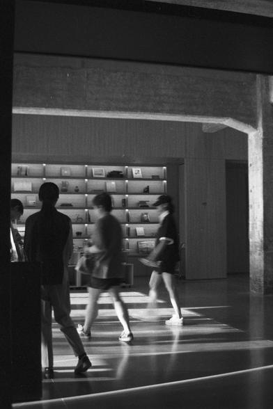 Ilford HP5 Plus 400 (FF)

English Alt Text:
A black-and-white photo of a modern indoor space with clean lines and minimalist design. Four people are walking or standing near a wall with built-in shelves filled with books and small objects. The floor is smooth and reflective, and the lighting casts soft shadows. Exposed concrete beams add an industrial touch. The scene feels like a gallery, museum, or bookstore, capturing quiet motion and thoughtful architecture.

中文替代文字：
这是一张黑白照片，展示一个现代室内空间，设计简…