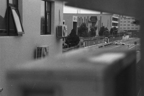 Ilford HP5 Plus 400 (FF)

English Alt Text:
A grainy black-and-white image taken from above, possibly through a window. It shows a street lined with institutional buildings, each with air conditioning units on the exterior. Cars are parked and moving along the road. Trees and streetlights dot the sidewalk, and a fence separates pedestrians from traffic. The photo has a vintage, film-like texture, evoking a quiet urban moment.

中文替代文本：
这是一张黑白照片，从高处俯拍，可能透过窗户拍摄。画面中是一条街道，两旁是带有外墙空调的机构建筑。路上有车辆停靠或行驶。人…