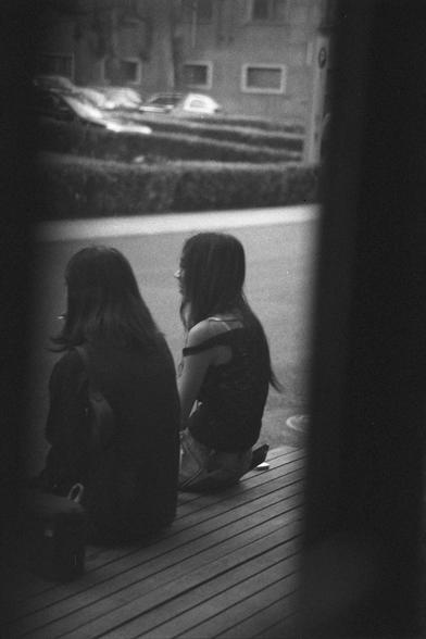 Ilford HP5 Plus 400 (FF)

English Alt Text:
A black-and-white photo framed by dark vertical edges, showing two people sitting side by side on a wooden surface outdoors. One wears a sleeveless top and shorts, the other a darker outfit. They face away from the camera, possibly talking or resting. Behind them are hedges, parked cars, and a building with windows. The scene feels intimate and reflective.

中文替代文本：
这是一张黑白照片，画面两侧有深色边框。两人并肩坐在户外的木质平台上，背对镜头。其中一人穿着无袖上衣和短裤，另一人穿着深色衣物。他们可能在交谈或静坐。背景中有绿篱、停放的车辆和…