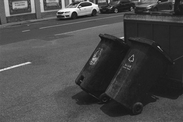 Ilford HP5 Plus 400 (FF)

English Alt Text:
A black-and-white photo of a gritty urban street. In the foreground, two large garbage bins lean against a dumpster—one marked with a recycling symbol and Chinese text. The bins are worn and stained. Behind them, cars move and park along the street, including a white sedan and a black SUV. Buildings line the road, with posters in Chinese on the walls. The scene captures the texture of everyday city life and waste management.

中文替代文本：
这是一张黑白照片，描绘了城市街道的…