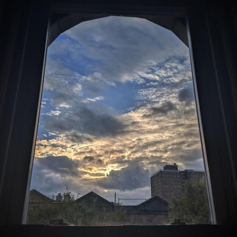 Looking through an arched window and hour after sunrise the blue sky is filled with complex layers of gray and white storm clouds. Across the middle of the sky is a band of backlit yellow clouds. Pointed roofs of Harlem brownstones are silhouetted across the street, and a taller apartment building can be seen in the distance. The tops of two trees are on the bottom and right.