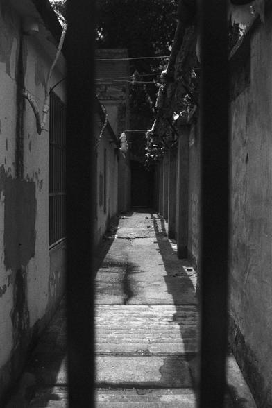 Ilford HP5 Plus 400 (FF)

English Alt Text:
A narrow alleyway between two aged buildings is viewed through vertical black bars in the foreground. The concrete path is cracked and stained. The walls are peeling, with exposed pipes and wires overhead. A barred window sits on the left wall. The alley leads into a darker area, evoking mystery. Sunlight casts sharp shadows, and the black-and-white palette emphasizes texture and decay.

中文替代文字：
透过前景中的黑色栏杆，可见两栋老旧建筑之间的狭窄小巷。混凝土地面布满裂痕与污渍，墙面斑驳脱落，上方管线交错。左侧…