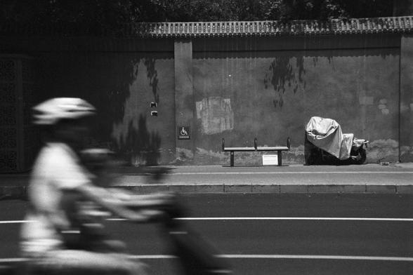 Ilford HP5 Plus 400 (FF)

English Alt Text:
Two blurred figures on bicycles or scooters speed past in the foreground, capturing motion. Behind them, a quiet urban wall features a handicapped-accessible sign, a bench with a small paper, and a covered object—possibly a scooter. Tree shadows fall across the wall. The contrast between movement and stillness is heightened by the monochrome tones.

中文替代文字：
前景中两位骑行者模糊掠过，展现速度与动感。背景是一面城市墙壁，设有残障通道标志、长椅和一张小纸条，旁边覆盖着一件物品，可能是电动车。树影洒落墙面，黑白色调强化了动与静的对比。