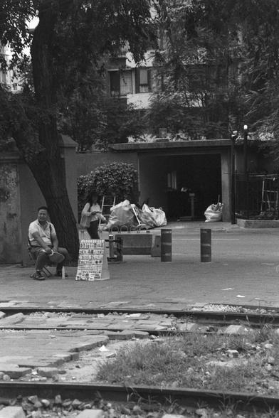 Ilford HP5 Plus 400 (FF)

English Alt Text:
Railway tracks cross a paved area in the foreground. Two people sit near a tree—one on a chair, the other beside a small stand with a signboard. A bench behind them holds large bags, possibly recyclables. A building with windows and an open garage is in the background. Bollards line the area, and tree shade softens the scene. The monochrome effect adds depth and quietness.

中文替代文字：
前景中铁轨穿过铺设区域。两人坐在树旁，一人坐椅上，另一人靠近带招牌的小摊。后方长椅上堆放着大袋子，可能是可回收物。背景是有窗户和敞开车库的建…