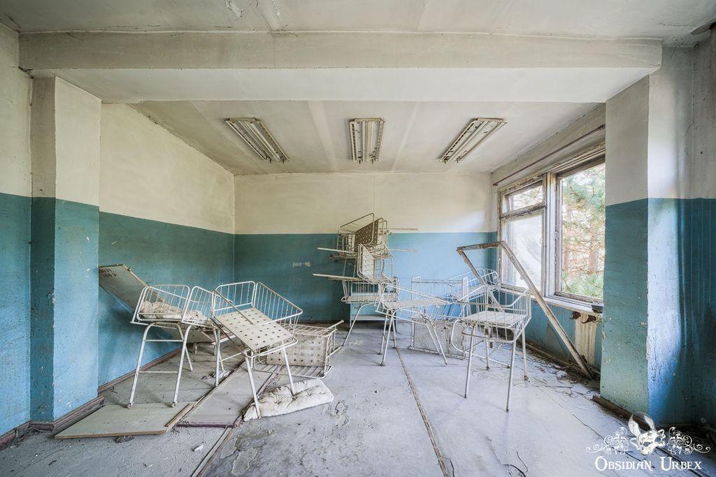 Abandoned room with peeling paint, broken windows, and several rusted, overturned metal hospital cribs scattered on the floor.