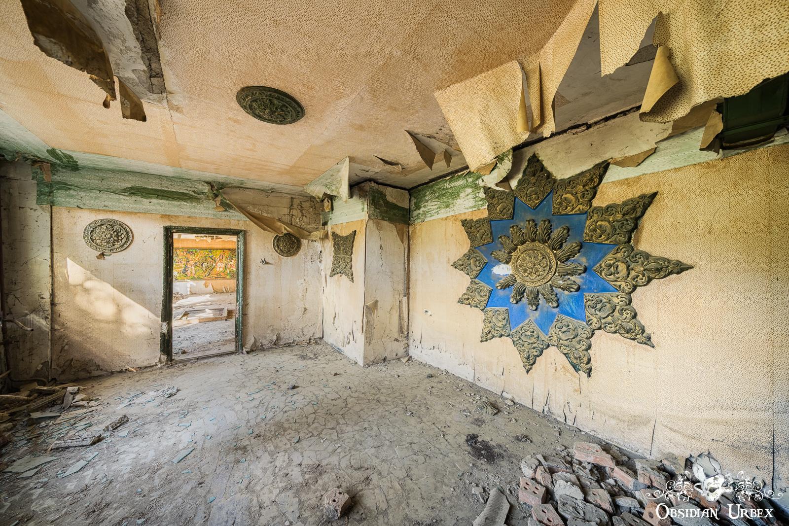 A decaying room with peeling ceiling, cracked floor, ornate blue and gold wall decorations, and an open doorway showing debris outside.