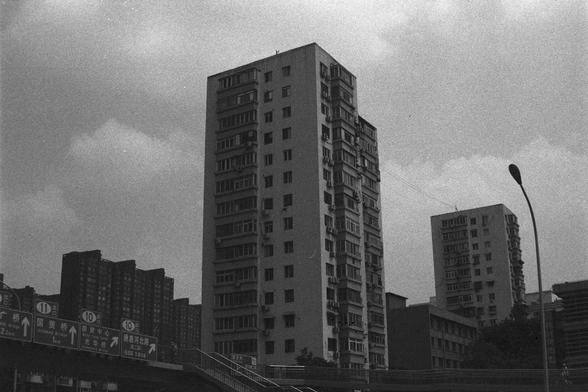 Ilford HP5 Plus 400 (FF)

English Alt Text:
A moody black-and-white photo of several tall residential buildings with balconies and windows. A streetlight stands in the foreground on the right. Road signs with Chinese characters and arrows are visible in the lower left, indicating a location in a Chinese city. The sky is cloudy, adding a somber tone. The image captures urban density and architectural repetition, evoking feelings of introspection and modern life.

中文替代文字：
这是一张黑白照片，画面中有几栋高大的住宅楼，带有…