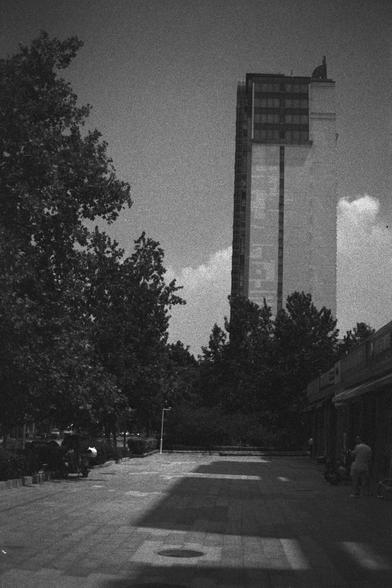 Ilford HP5 Plus 400 (FF)

English Alt Text:
A black-and-white image of a sleek high-rise building in the background, surrounded by trees. In the foreground, a paved pedestrian walkway is shaded by trees, with a few people sitting or walking. Bicycles are parked on the right side. The building features glass and concrete, reflecting modern architecture. The scene feels calm and urban, with natural light casting shadows that add depth and texture.

中文替代文字：
这是一张黑白照片，背景是一栋现代高层建筑，外观由玻璃和混凝土构成，周围环绕着树木…