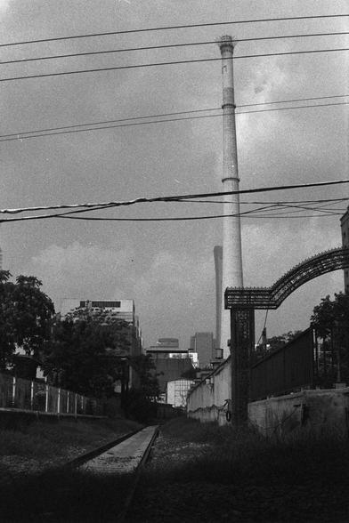 Ilford HP5 Plus 400 (FF)

English Alt Text:
A black-and-white photo of an industrial area featuring a narrow railway track leading into the distance. A tall smokestack dominates the center-right, with another visible in the background. Overhead power lines cross the top. On the right, a decorative metal archway is attached to a wall. Trees line the left side, contrasting with the industrial buildings. The composition blends gritty textures with natural elements, creating a nostalgic and layered…