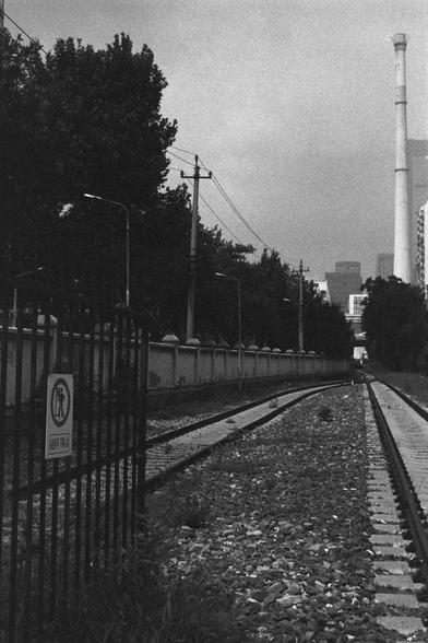 Ilford HP5 Plus 400 (FF)

English Alt Text:
A black-and-white photo showing a straight railway track stretching into the distance. On the left, a tall metal fence bears a “KEEP WALK” sign with a symbol prohibiting pedestrian access. Behind the fence are trees and utility poles with overhead wires. A concrete wall with evenly spaced pillars runs parallel to the track. In the hazy background, tall buildings and a smokestack rise, suggesting an industrial urban setting. The composition uses strong…