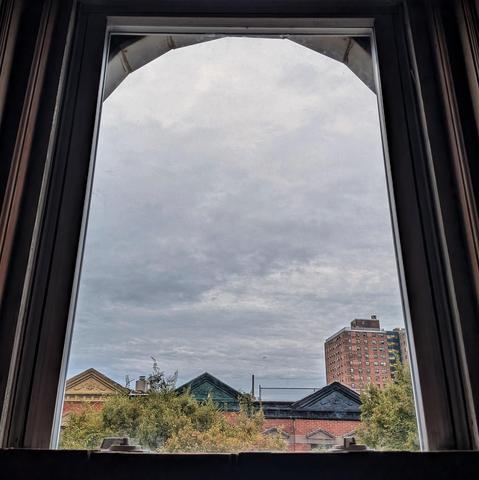 Looking through an arched window half an hour after sunrise the sky is full of gray rain clouds. Pointed roofs of Harlem brownstones with red brickwork are across the street, and a taller apartment building can be seen in the distance. The green tops of two trees are on the bottom and right.