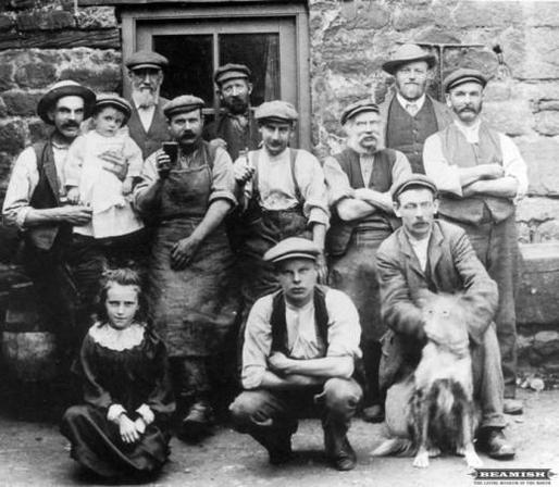 Group photo from late 19th century or early 20th century. A row of eight men of varying heights standing in front of a brick wall with a casement window behind them. Two men are squatting or kneeling in front of them in center and at right, with a dog sitting in front at the right. The dog, for some reason, appears to have googly eyes. A young, possibly pre-teen girl is sitting on the ground at front left. She’s wearing a long dark dress with a white lace collar. All of the men are wearing hats…