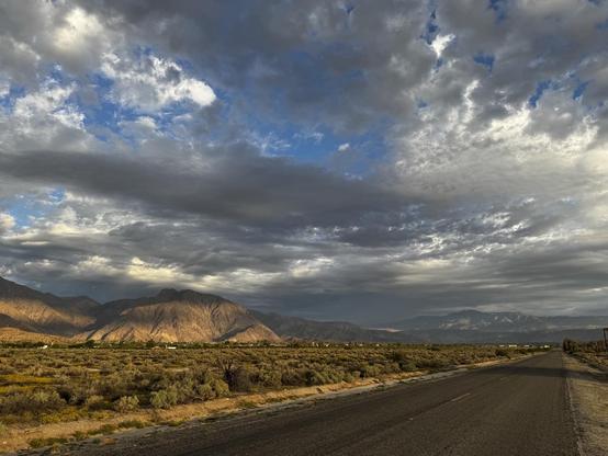 iPhone photo of a road stretching into the distance, flanked by desert grasses, scrub, scattered vegetation. Majestic mountains rise in the background under a dramatic sky filled with grey clouds, creating a serene and expansive landscape.