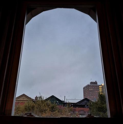 Looking through an arched window ten minutes after sunrise the sky is full of grey rain clouds. There are a few raindrops on the glass. Pointed roofs of Harlem brownstones with red brickwork are across the street, and a taller apartment buildifng can be seen in the distance. The green tops of two trees are on the bottom and right.