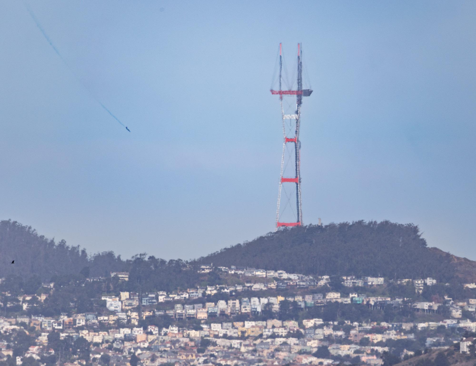 A photo of San Francisco's Sutro Tower with a jet arcing downward through the sky over hills with lines of houses.