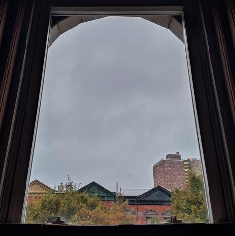 Looking through an arched window at the moment of sunrise the sky is full of gray clouds. Pointed roofs of Harlem brownstones with red brickwork are across the street, and a taller apartment buildifng can be seen in the distance. The green tops of two trees are on the bottom and right.