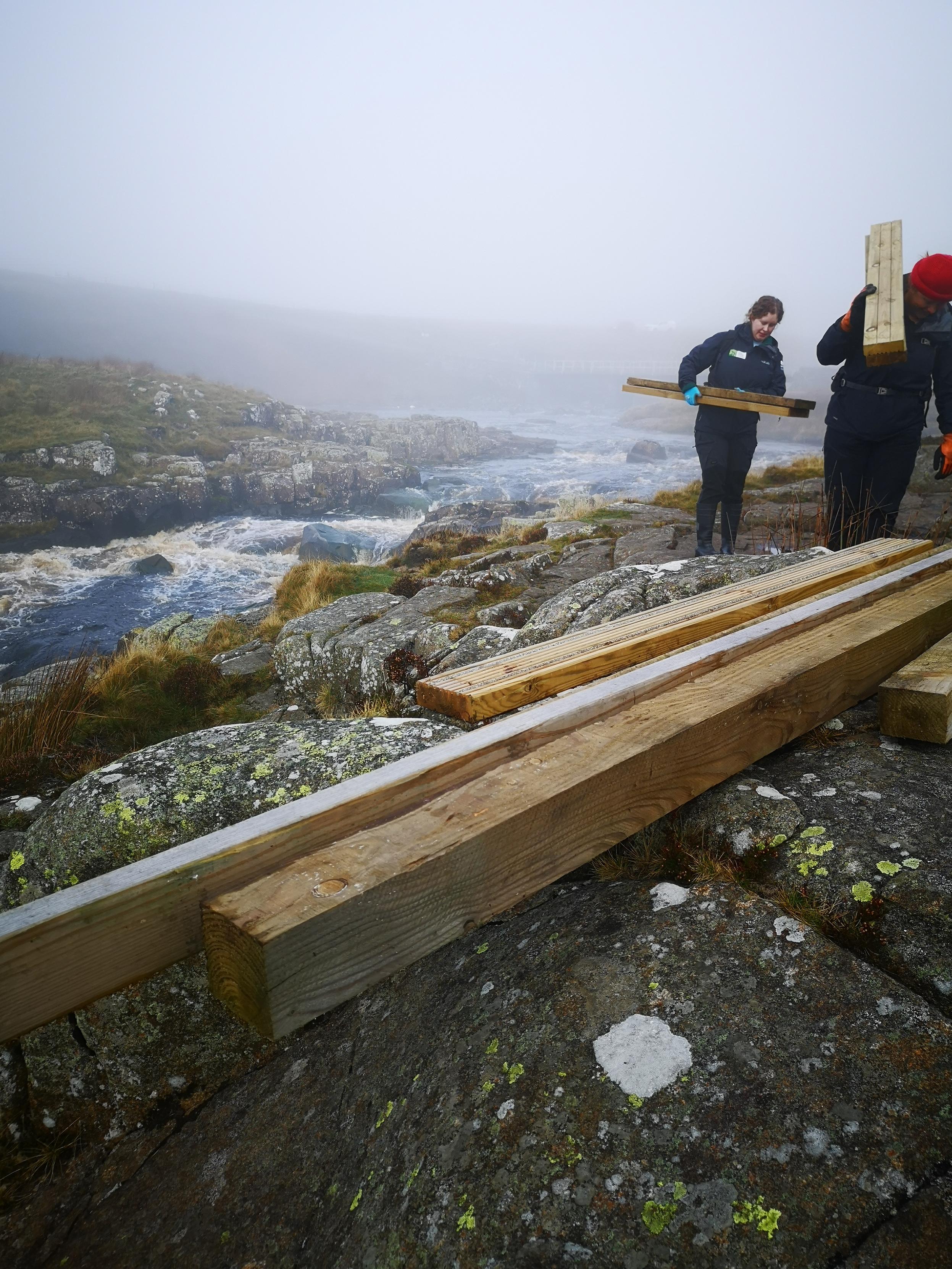 It's a foggy day by a stream rushing down from Cow Green reservoir. There are pieces of timber on the ground and two women in dark clothing also carrying timber behind those. The ground is rocky with rough grasses.