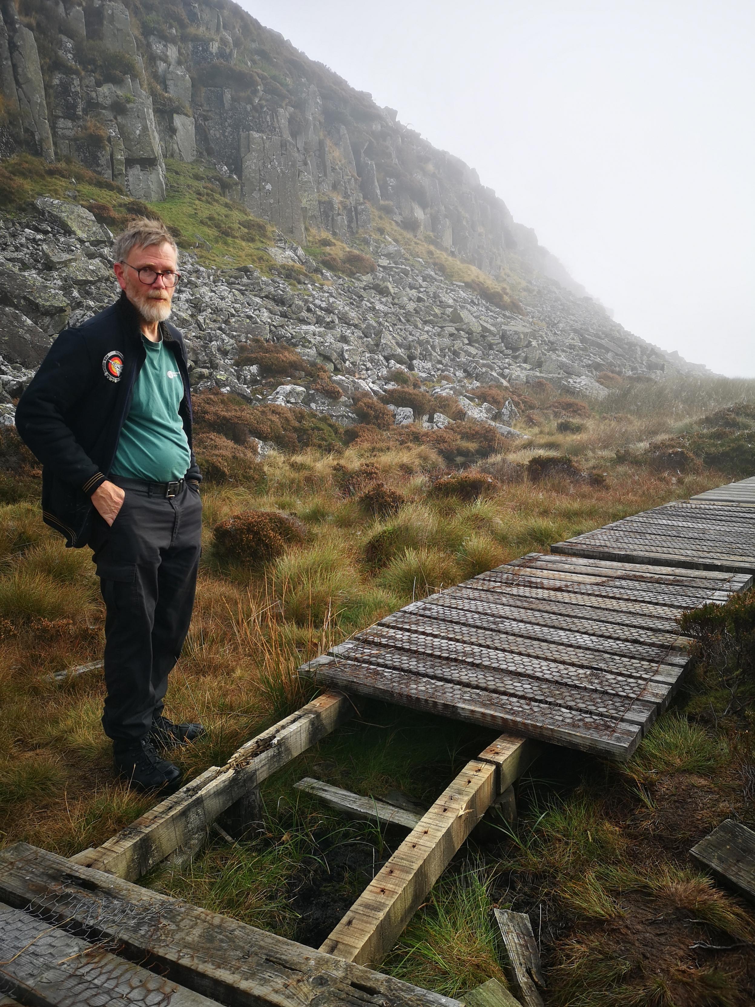 A fellow volunteer stands beside a length of wooden boardwalk which has some of the timber treads missing, exposing the supporting timbers. Behind is the grey rock face of the Whin Sill, a dolerite intrusion that outcrops in many places in North Pennines. The weather is still but misty.
