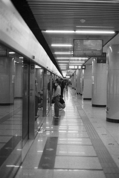 Kentmere Kentmere PAN 400 (FF)

English Alt Text:
A clean, modern subway station platform with glass barriers separating passengers from the tracks. People stand or sit near the glass, waiting for the train. Overhead, striped ceiling panels with embedded lights stretch across the space. Hanging signs display information in Chinese characters. The floor is tiled with tactile paving for visually impaired navigation. Cylindrical columns support the structure, and the overall atmosphere is orderly …