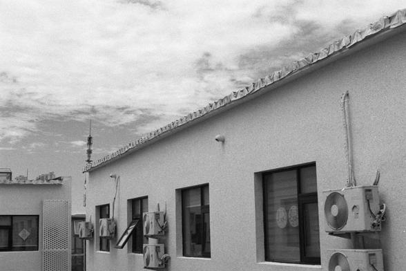 Kentmere Kentmere PAN 400 (FF)

English Alt Text:
A black-and-white photo of a multi-story building with numerous air conditioning units mounted beneath each window. The building has a flat roof with a corrugated edge. Above it, the sky is partly cloudy. In the background, a tall antenna or communication tower rises, suggesting an urban or semi-urban location. The image has a vintage, documentary feel, emphasizing the industrial and functional aspects of the architecture.

中文替代文字：
这是一张黑白照片，展示了一…