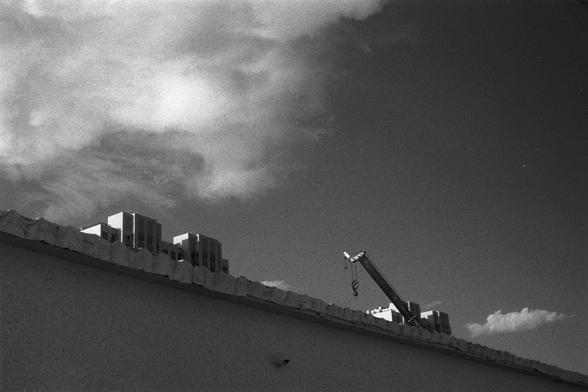 Kentmere Kentmere PAN 400 (FF)

English Alt Text:
A black-and-white image showing the top of a building with a corrugated roof. Several rectangular HVAC units are installed on the roof. A crane arm extends upward from the right side, with a hook hanging down. The sky is partly cloudy, with dramatic contrasts between light and dark clouds. The composition highlights industrial elements against a natural sky.

中文替代文字：
这是一张黑白照片，画面为一栋建筑的屋顶，屋顶呈波纹状，上面安装了多个矩形的空调设备。右侧有一只起重机臂向上延伸，吊钩悬挂在空中。天空中云层交错，明暗对比强烈，…