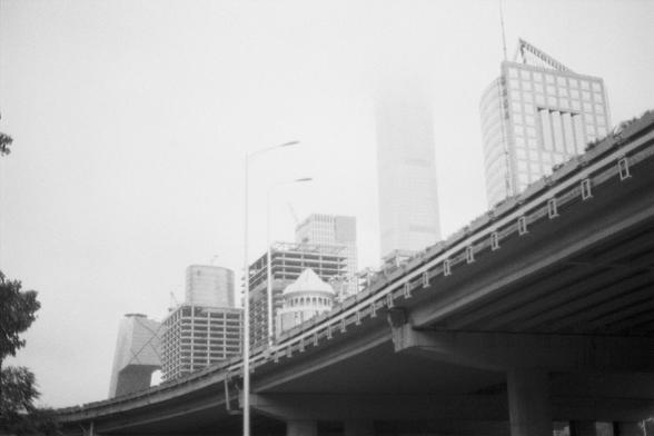 Kentmere Kentmere PAN 400 (FF)

English Alt Text:
A grayscale cityscape featuring modern high-rise buildings with varied architectural styles—some cylindrical, others rectangular with glass facades. An elevated highway cuts diagonally across the foreground, supported by concrete pillars. Streetlights line the road, and a tree sits on the left. Fog or low clouds partially obscure one building, adding a moody, atmospheric tone.

中文替代文字：
这是一幅灰度城市景观图，展示了多座现代高楼，建筑风格各异，有圆柱形，也有玻璃幕墙的矩形结构。前景中，一条高架公路斜穿画面…