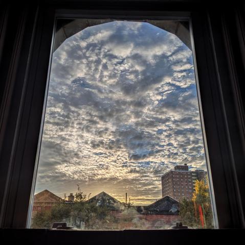 Looking through an arched window half an hour after sunrise the blue sky is full of spidery grey and white clouds, and there is a yellow haze above the horizon. Pointed roofs of Harlem brownstones with red brickwork are across the street, and a taller apartment buildifng can be seen in the distance. The green tops of two trees are on the bottom and right. The window is grimy on the bottom.