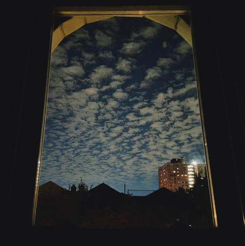Looking through an arched window a few hours before sunrise the dark blue sky is filled with rows of small white clouds. Pointed roofs of Harlem brownstones are silhouetted across the street, and a taller apartment building can be seen in the distance.