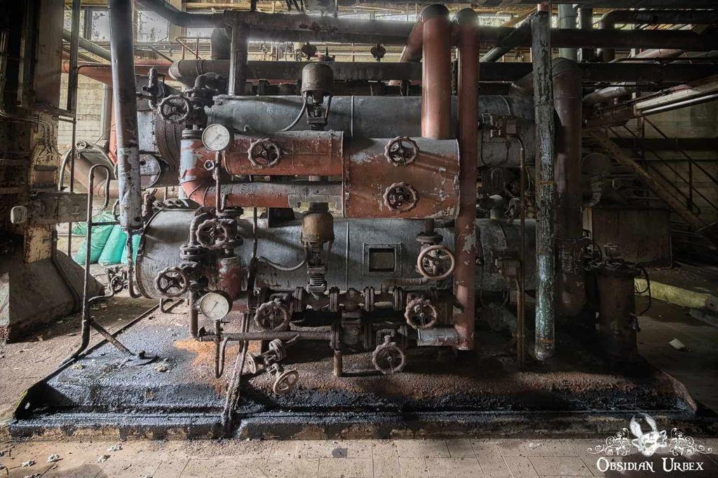A large, rusted industrial machine with pipes, gauges, and valves in an abandoned facility, showing signs of age and decay.