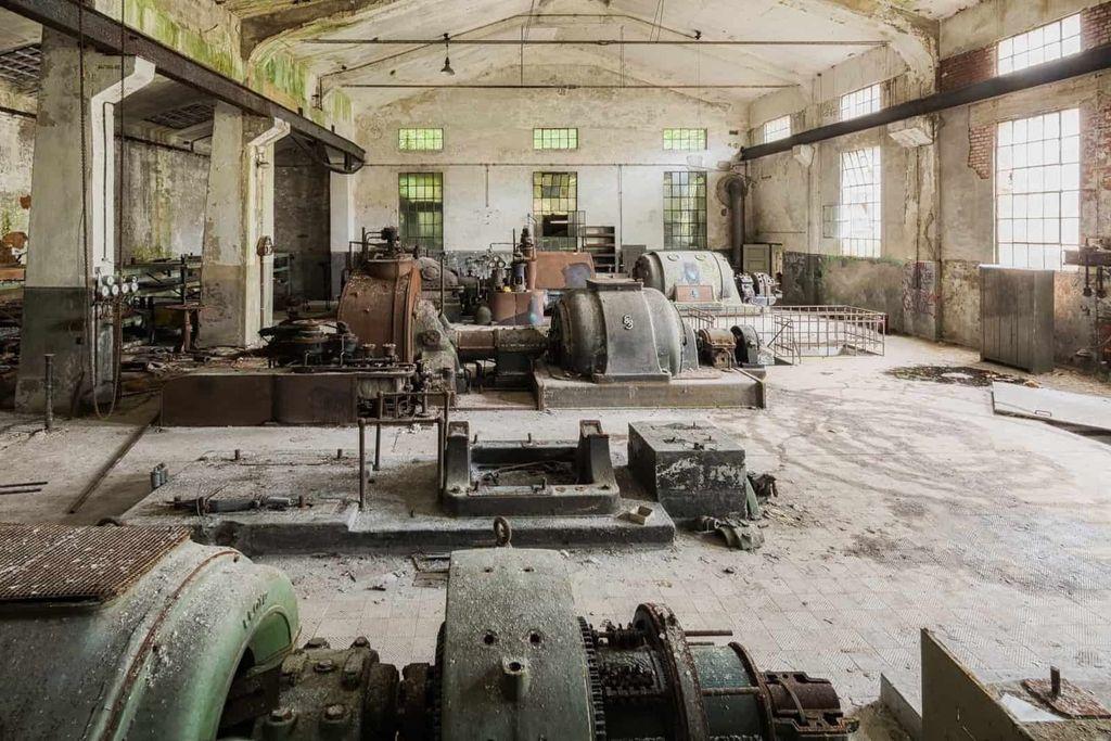 An abandoned industrial facility with old, rusted machinery and peeling paint, illuminated by natural light from large windows.